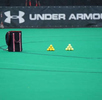 A sports field features a black and pink sports bag, two pyramids of yellow balls, and a portion of a person in dark athletic gear. The background displays a banner with the Under Armour logo.