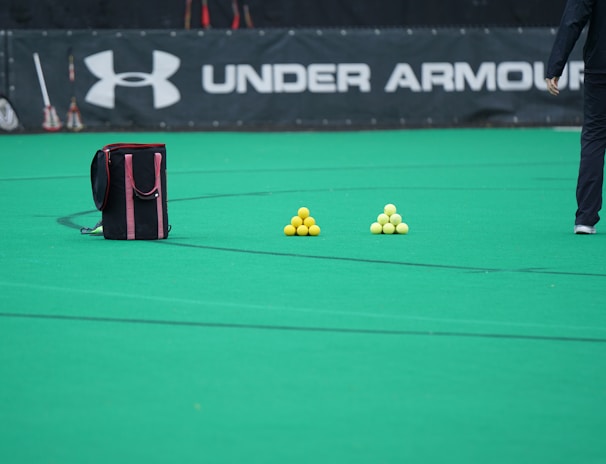 A sports field features a black and pink sports bag, two pyramids of yellow balls, and a portion of a person in dark athletic gear. The background displays a banner with the Under Armour logo.