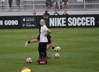 A person in sports attire stands on a soccer field, surrounded by several soccer balls and a carrier with bottles. The field is bordered by advertisements for Nike Soccer and app stores. Several people sit in bleachers in the background.