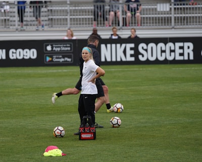 A person in sports attire stands on a soccer field, surrounded by several soccer balls and a carrier with bottles. The field is bordered by advertisements for Nike Soccer and app stores. Several people sit in bleachers in the background.