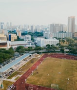 An aerial view of a sports complex with a running track surrounding a grassy field, set against the background of a cityscape. The city's skyline features a mix of tall residential buildings and shorter commercial structures. There are trees and green areas surrounding the sports facility.