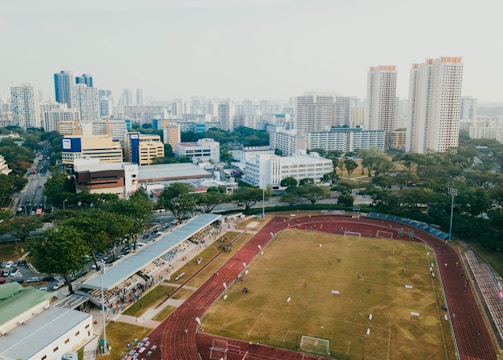 An aerial view of a sports complex with a running track surrounding a grassy field, set against the background of a cityscape. The city's skyline features a mix of tall residential buildings and shorter commercial structures. There are trees and green areas surrounding the sports facility.