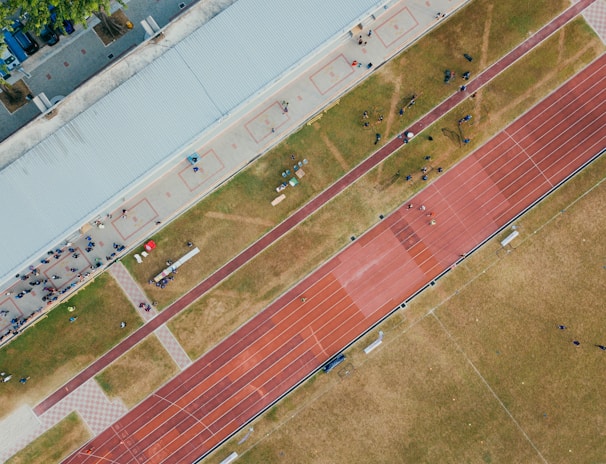 An aerial view of a sports complex featuring a running track and a grassy field. The tracks are red and situated next to a large building. There are several people scattered across the area, some on the track and others near the building. The surroundings include trees and geometric pathways, adding structure to the scene.