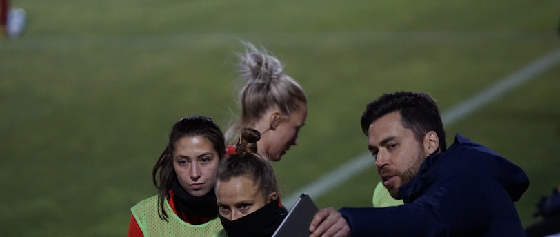 A group of individuals on a sports field are wearing green vests over red attire. One person is holding a tablet and seems to be discussing or showing something to the others. The atmosphere suggests a sports event, likely soccer, indicated by their clothing and the setting. The background is a grassy field.