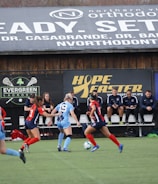 Players engage in a competitive soccer game on a field while others are seated on the bench watching attentively. The action is taking place in front of a wooden structure displaying multiple advertisements, including orthodontics, lacrosse, and field hockey.