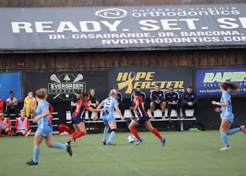 Players engage in a competitive soccer game on a field while others are seated on the bench watching attentively. The action is taking place in front of a wooden structure displaying multiple advertisements, including orthodontics, lacrosse, and field hockey.