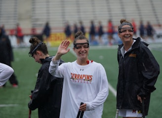 Several athletes in sports attire are on an outdoor sports field, interacting and smiling. One individual in the foreground waves with a friendly expression, while another stands beside her, also smiling. They wear protective goggles and gear associated with lacrosse. The background features indistinct figures in dark clothing against a backdrop of stadium seating.