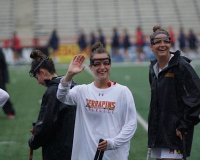 Several athletes in sports attire are on an outdoor sports field, interacting and smiling. One individual in the foreground waves with a friendly expression, while another stands beside her, also smiling. They wear protective goggles and gear associated with lacrosse. The background features indistinct figures in dark clothing against a backdrop of stadium seating.