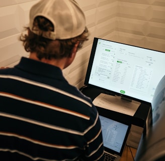 A person with a striped shirt and a cap is standing in front of a computer screen displaying data and analytics. Another laptop is open on a desk below the monitor, and a large metallic tank with a pressure gauge is nearby.