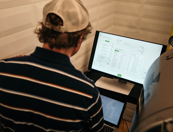 A person with a striped shirt and a cap is standing in front of a computer screen displaying data and analytics. Another laptop is open on a desk below the monitor, and a large metallic tank with a pressure gauge is nearby.