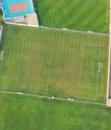 An aerial view of a sports complex featuring multiple green grass fields, including football pitches with marked goalposts. Adjacent to the fields is a blue-roofed building and a smaller area with a tennis or basketball court. Surrounding the complex, there's a road lined with trees and a dirt area with sparse vegetation.