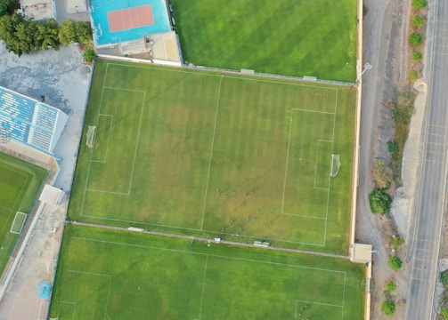 An aerial view of a sports complex featuring multiple green grass fields, including football pitches with marked goalposts. Adjacent to the fields is a blue-roofed building and a smaller area with a tennis or basketball court. Surrounding the complex, there's a road lined with trees and a dirt area with sparse vegetation.