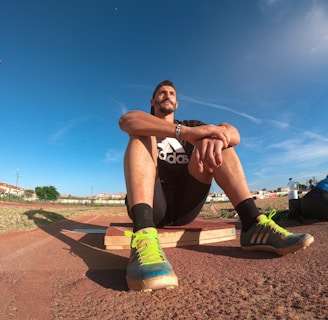 A person wearing a black athletic outfit is seated on a wooden platform in an outdoor sports field. The individual is relaxed, with arms resting on knees. The field is expansive with a clear blue sky and scattered clouds above. A brightly colored running shoe and athletic gear are visible, along with a water bottle and a blue backpack placed beside the person. Lampposts and residential buildings can be seen in the background.
