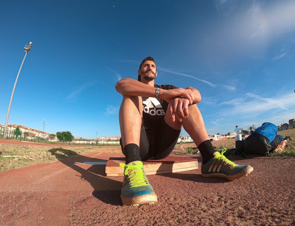 A person wearing a black athletic outfit is seated on a wooden platform in an outdoor sports field. The individual is relaxed, with arms resting on knees. The field is expansive with a clear blue sky and scattered clouds above. A brightly colored running shoe and athletic gear are visible, along with a water bottle and a blue backpack placed beside the person. Lampposts and residential buildings can be seen in the background.