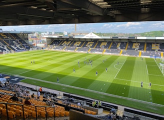 A sports stadium with a well-maintained grass field is depicted. Players are spread across the field, engaging in warm-up activities. The stands are partially filled with spectators, and the stadium is surrounded by urban buildings under a partly cloudy sky.