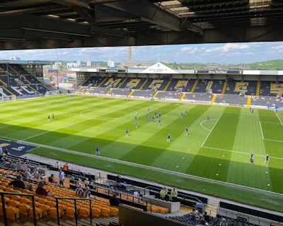 A sports stadium with a well-maintained grass field is depicted. Players are spread across the field, engaging in warm-up activities. The stands are partially filled with spectators, and the stadium is surrounded by urban buildings under a partly cloudy sky.