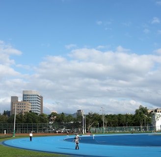 A bright outdoor sports complex features a blue running track, surrounded by green grass and trees. Several people are visible, engaged in activities on the track. In the background, urban high-rise buildings rise against a partly cloudy sky.