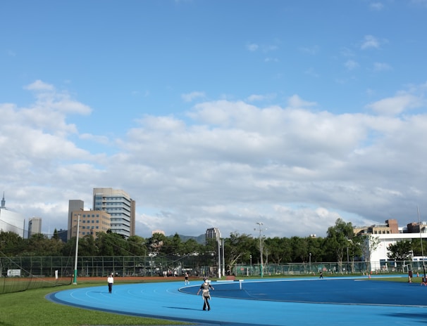 A bright outdoor sports complex features a blue running track, surrounded by green grass and trees. Several people are visible, engaged in activities on the track. In the background, urban high-rise buildings rise against a partly cloudy sky.