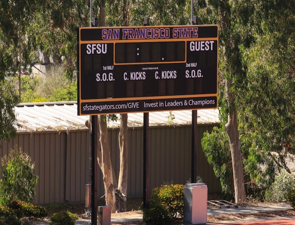 An outdoor scoreboard for San Francisco State is seen among trees and greenery. The board displays sections for shots on goal and corner kicks, with areas for both home and guest teams. It is situated near a running track with a building in the background.