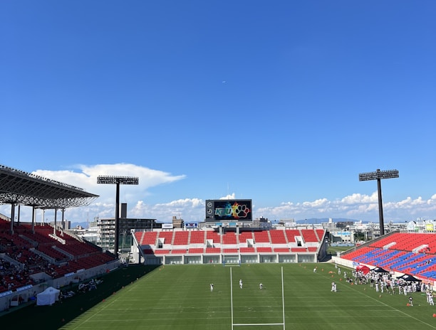 A large sports stadium with red and blue seating overlooks a green field with a goalpost at the center. A few people are scattered across the field, with some tents and a gathering of individuals on the right side. The sky is clear and blue with some clouds visible in the distance. A large screen or scoreboard is situated at the far end of the field.